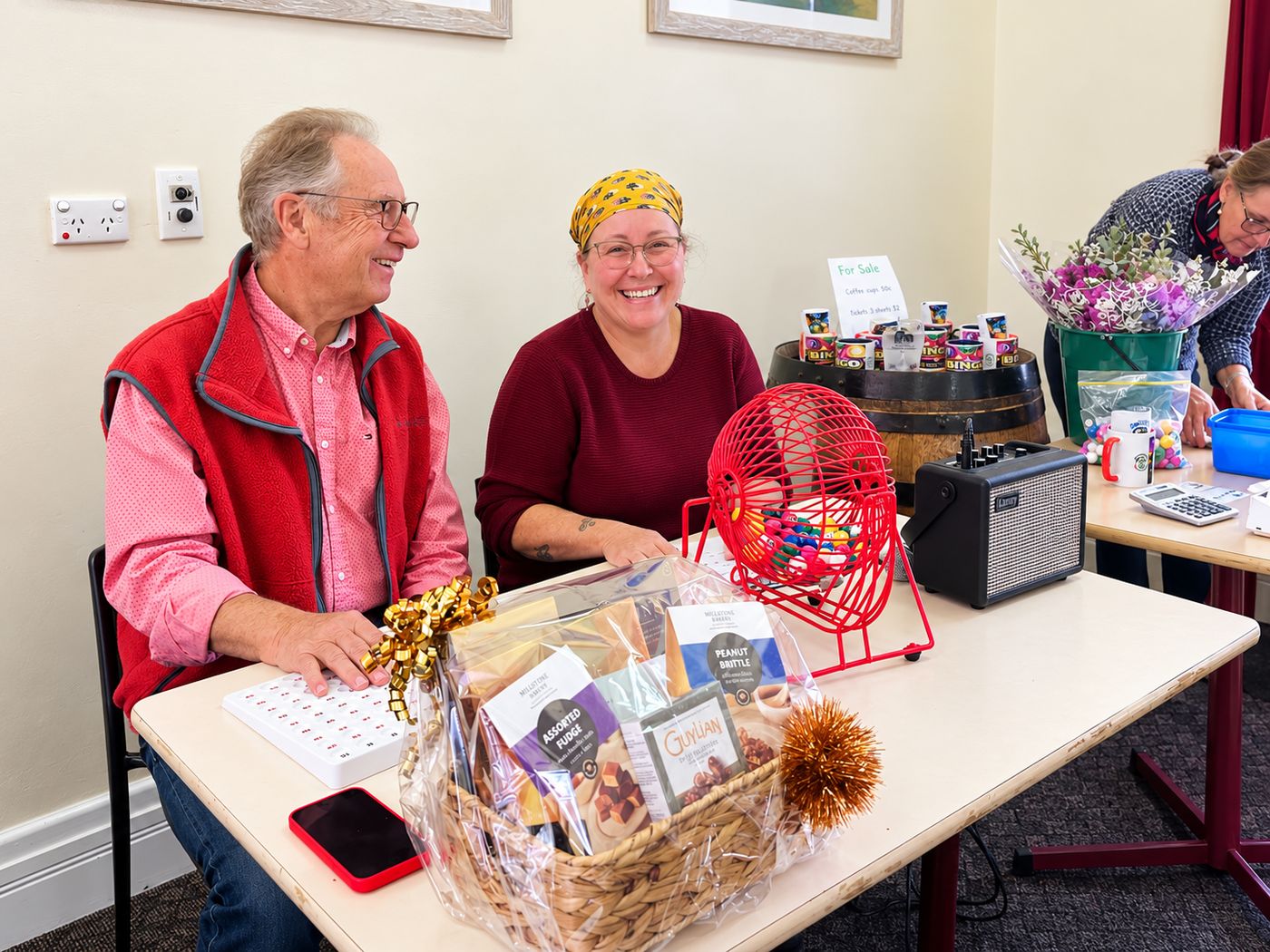 Two smiling volunteers at the bingo table with the ball cage and a prize hamper